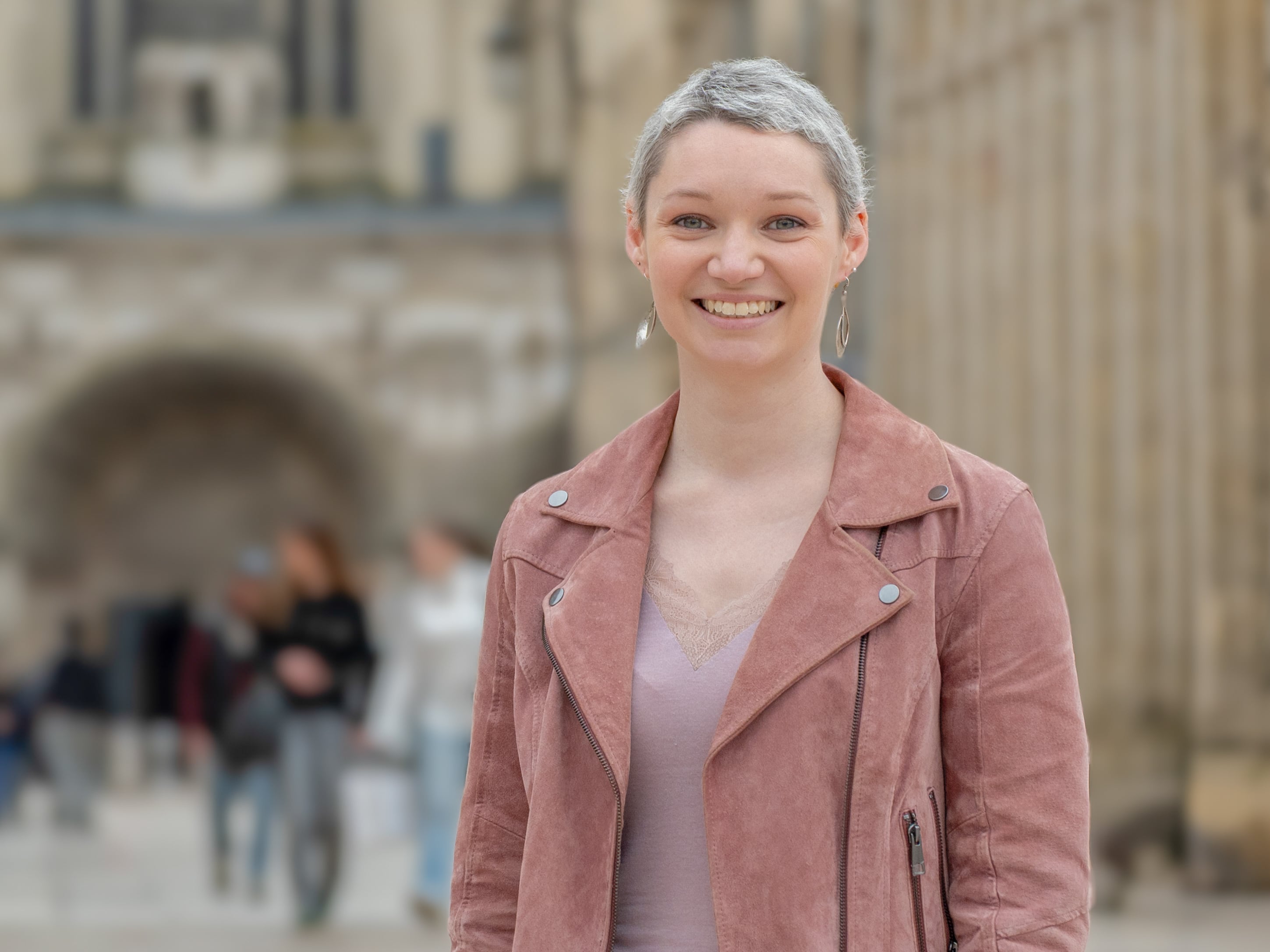 Elise is standing in front of a blurred city background with a smile and a pink jacket.