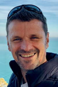 Thierry is smiling in front of a blue background. Sunglasses are resting on the top of his head.